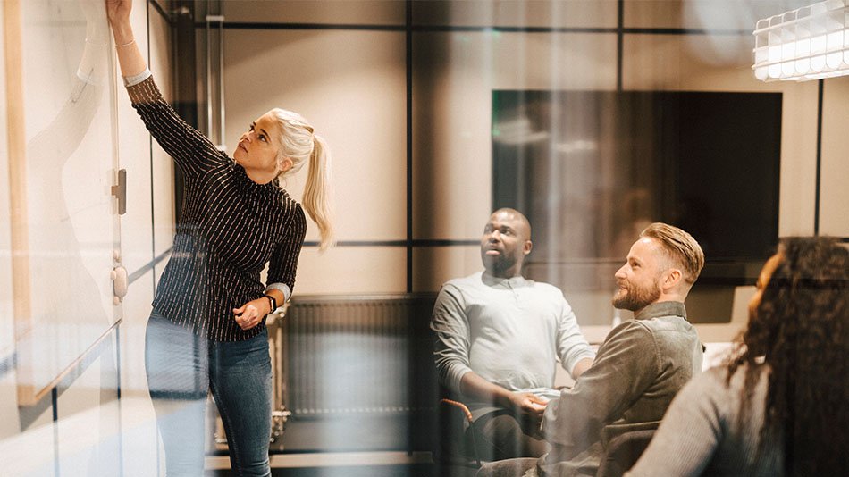 A meeting room. One woman is standing, writing something on the whiteboard. Three people sitting and listening to the the woman.
